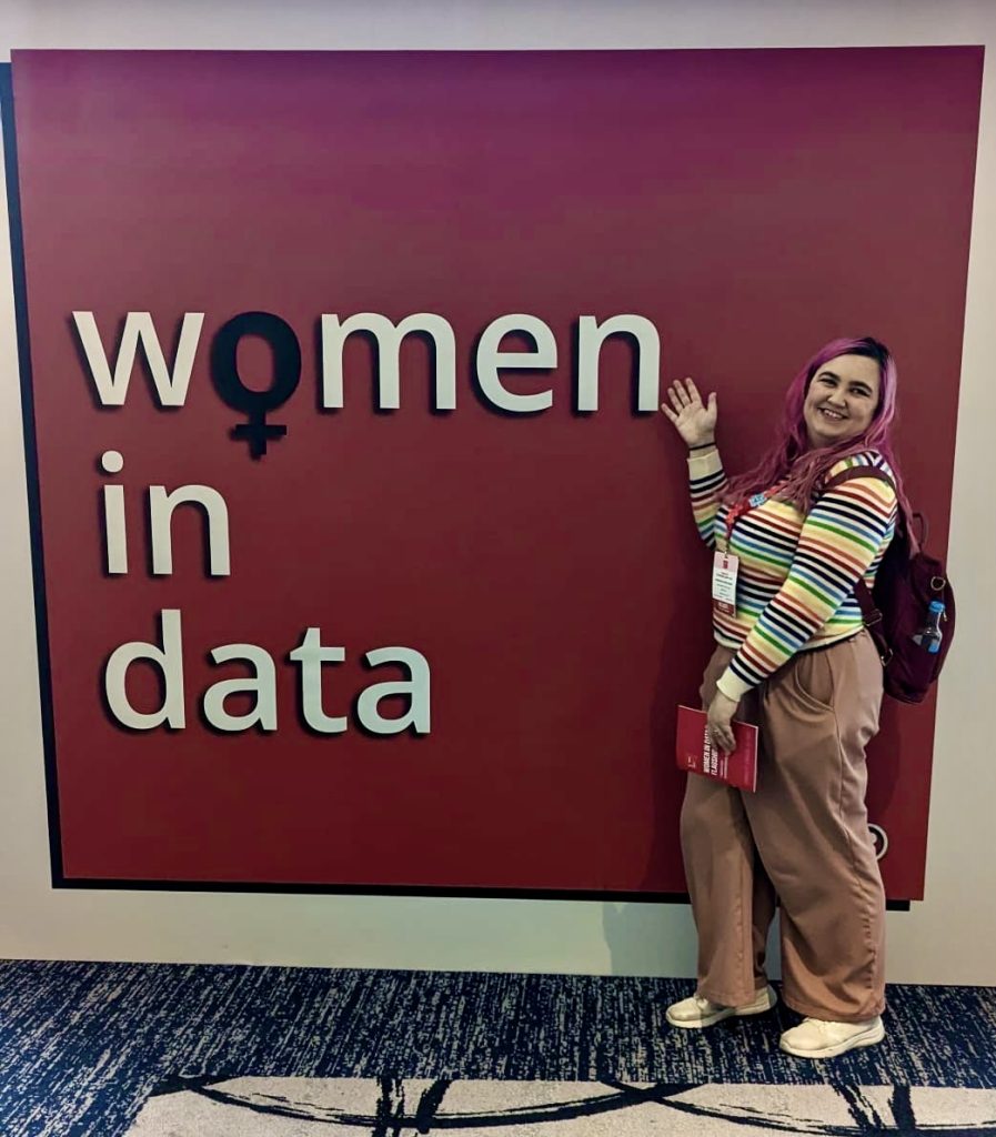 Georgiana posing in front of a promotional poster for the Women in Data conference 