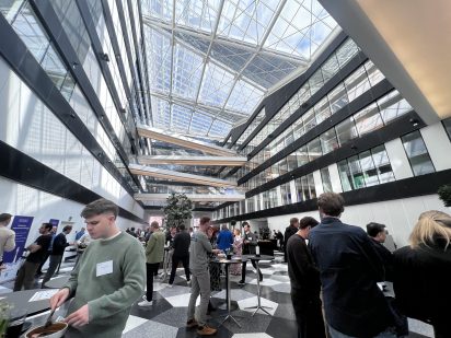 A bright, spacious room with a glass roof filled with natural light. Conference attendees are chatting and mingling during the lunch break at Cboost.