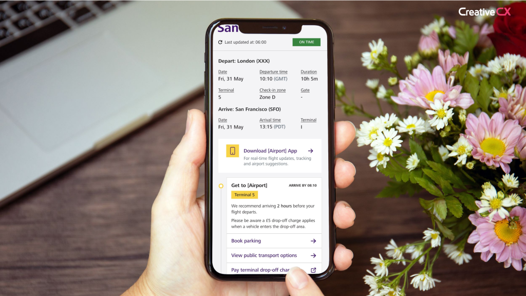 A person holds a smartphone displaying flight information for a flight to San Francisco. The screen shows departure from London on Friday, 31 May at 10:10 GMT and arrival in San Francisco at 13:15 PDT. The flight duration is 10 hours and 5 minutes. The app also includes check-in and terminal details, along with a prompt to download an airport app. In the background, there's a laptop and a bouquet of pink and white flowers on a wooden table. The Creative CX logo is visible in the top right corner