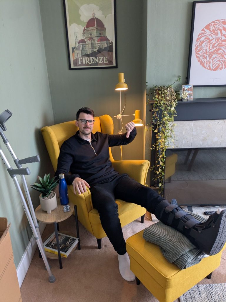 Andy (author of this post) with a broken ankle sits in a yellow armchair with his leg in a medical boot resting on a footstool. Crutches lean against the wall beside him. The image reflects the lived experience of temporary accessibility challenges.