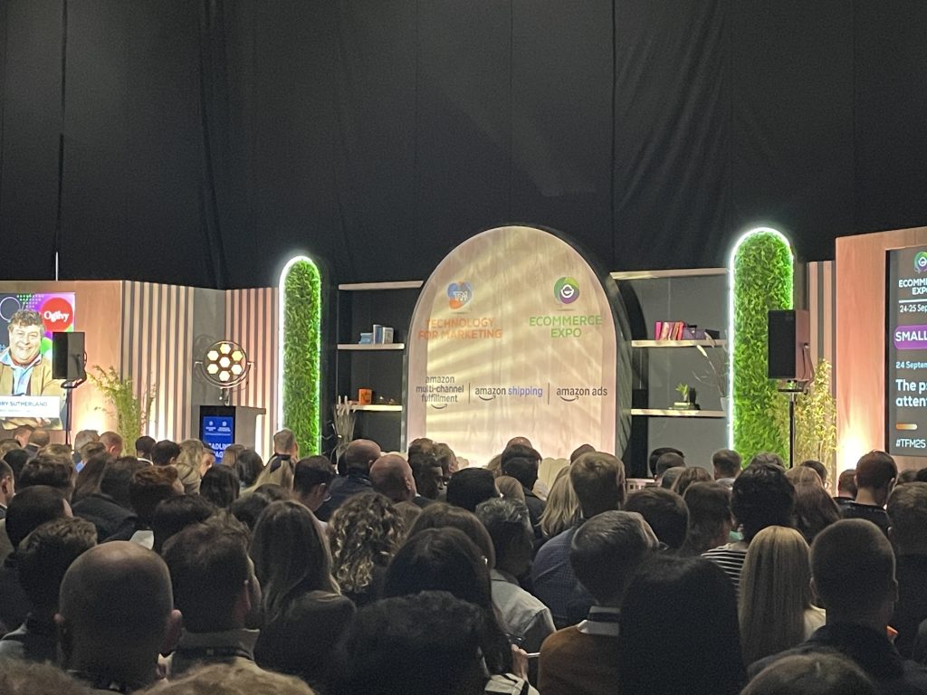 Large audience seated and watching a keynote presentation on a well-lit stage at eCommerce Expo 2025, with branded displays and illuminated greenery in the background.