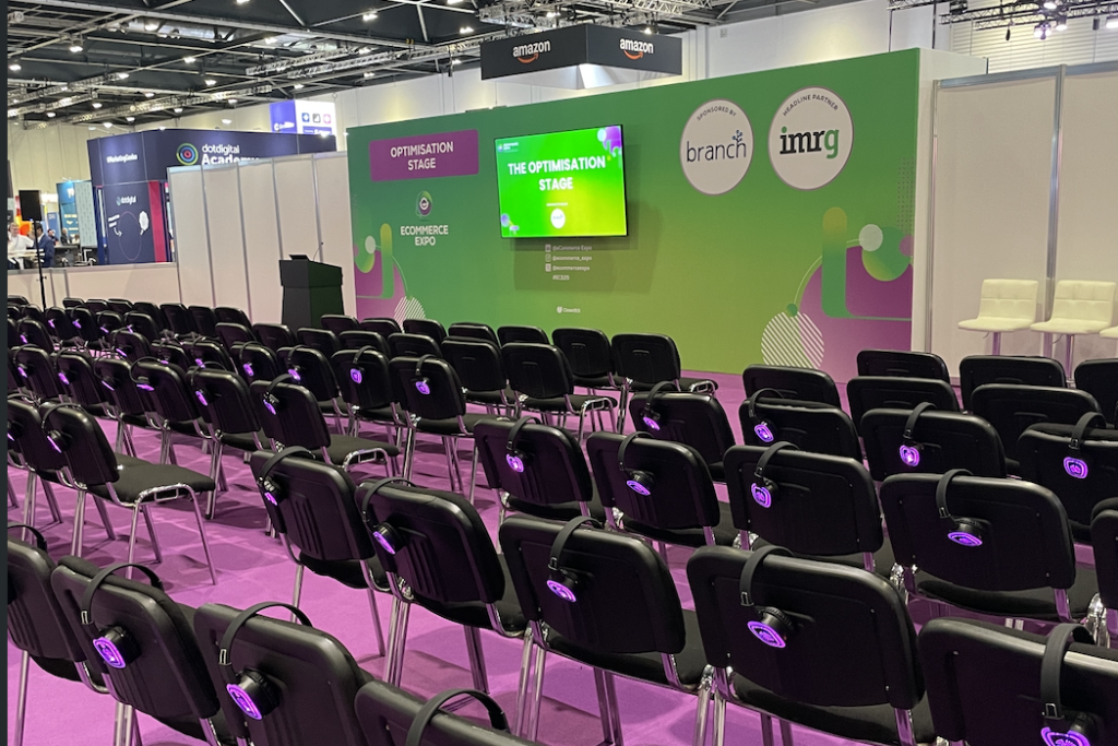 Rows of chairs with headphones set up in front of the Optimisation Stage at eCommerce Expo 2025, featuring a green and purple backdrop with sponsor logos.