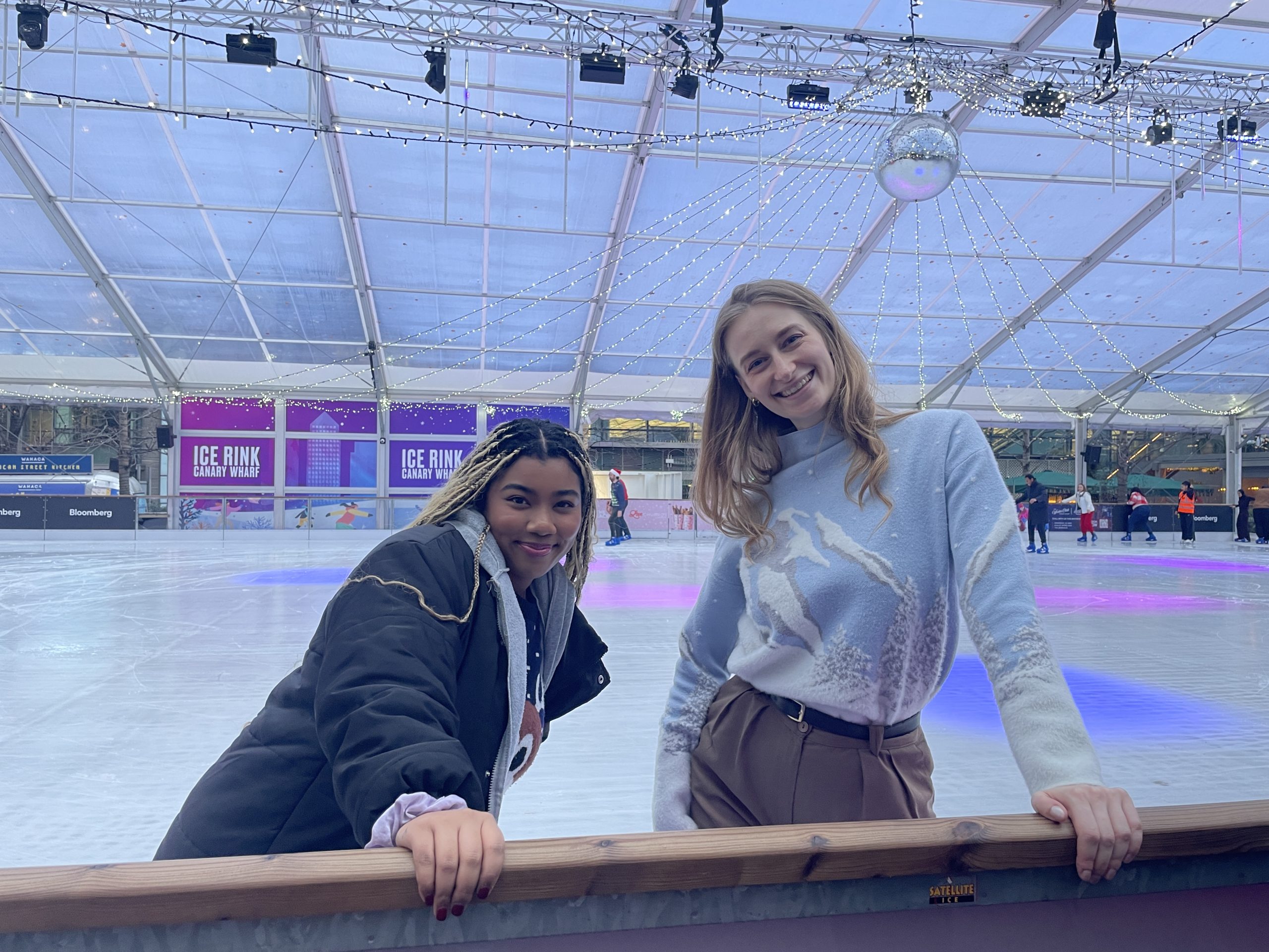 A photo of two Creative CX team members smiling at the edge of an indoor ice rink, with festive lights overhead and skaters in the background.