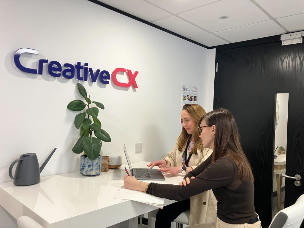 Two colleagues sit at a desk in an office, reviewing work on a laptop and taking notes. A “Creative CX” sign is displayed on the wall behind them, with a plant and workspace items visible, suggesting a collaborative working session.