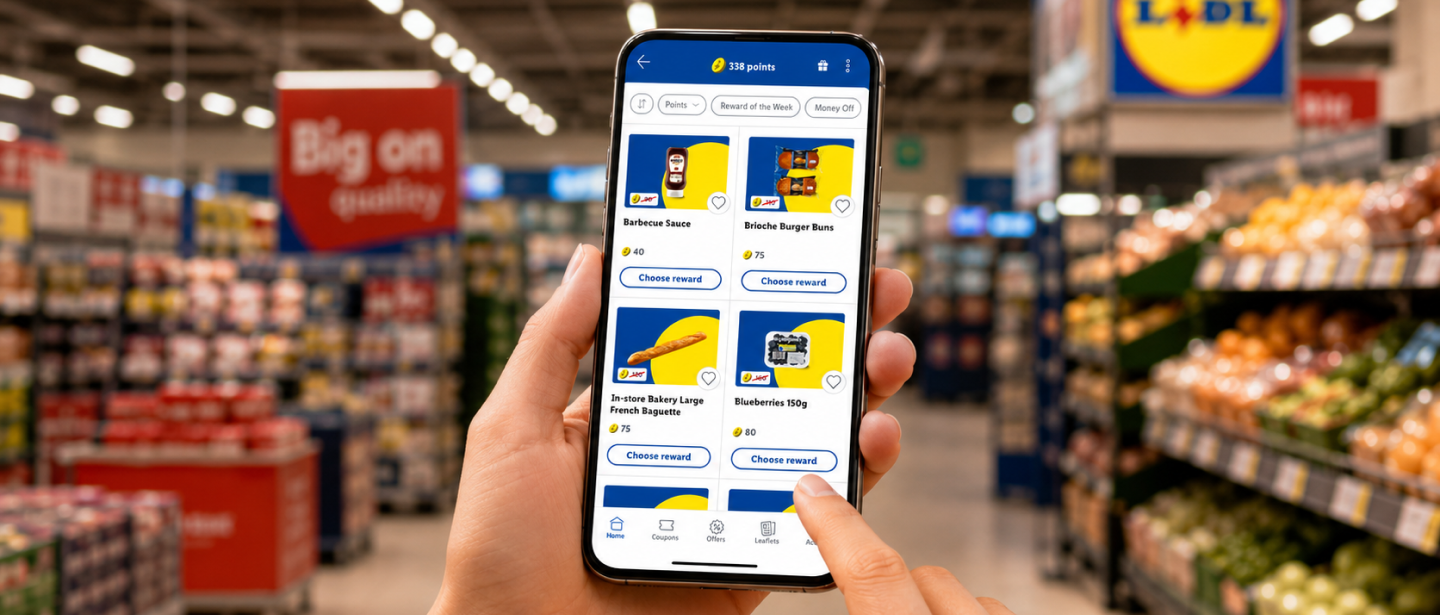 A close-up shot of a person’s hands holding a smartphone inside a brightly lit Lidl supermarket aisle. The phone screen displays the Lidl Plus loyalty app, showing 338 points at the top and a grid of grocery rewards available for selection, including barbecue sauce, brioche burger buns, a French baguette, and blueberries. The background shows blurred supermarket shelves stocked with products and fresh produce.