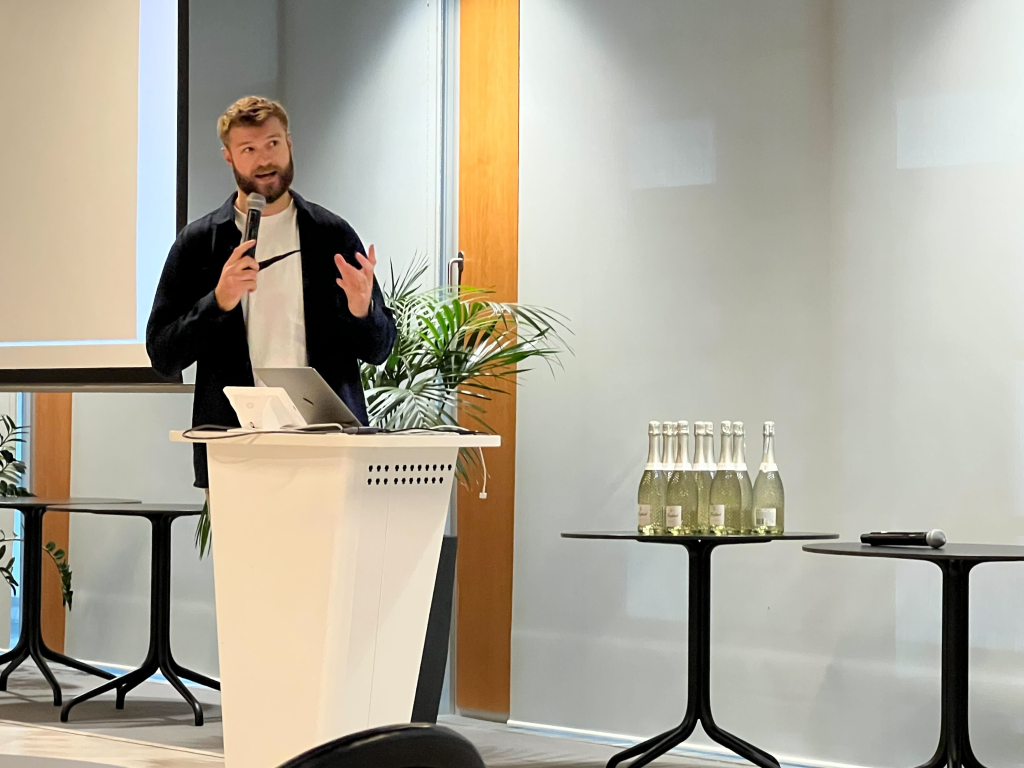Paul Wainwright, Senior Strategist at Creative CX, presenting during a YOOX Net-A-Porter Group hackathon. Standing behind a podium with a microphone and laptop, he shares ideas and insights with attendees in a modern conference space.