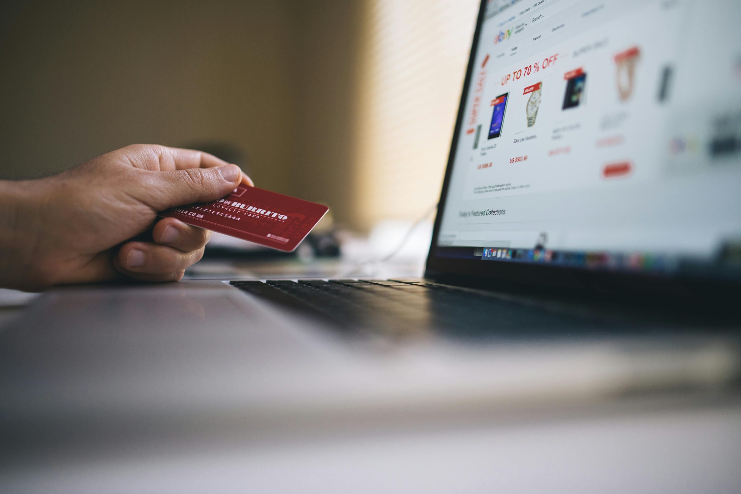 A person holds a red credit card in front of a laptop while shopping online. The laptop screen displays an e-commerce website with discounted products, and the image is softly focused to emphasise the online payment process.