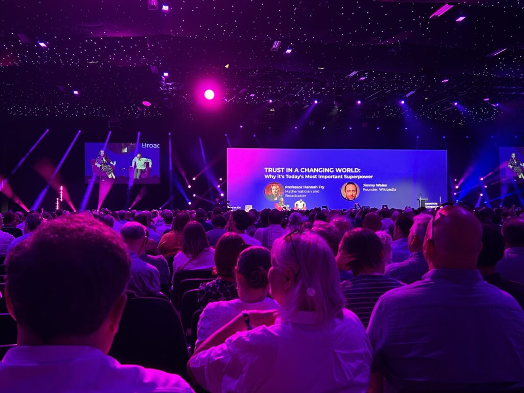 Audience view of a keynote session at the Octopus Energy Tech Summit. On stage, Professor Hannah Fry and Wikipedia founder Jimmy Wales take part in a discussion titled "Trust in a Changing World: Why it's Today's Most Important Superpower", with the presentation displayed on a large screen under purple stage lighting.