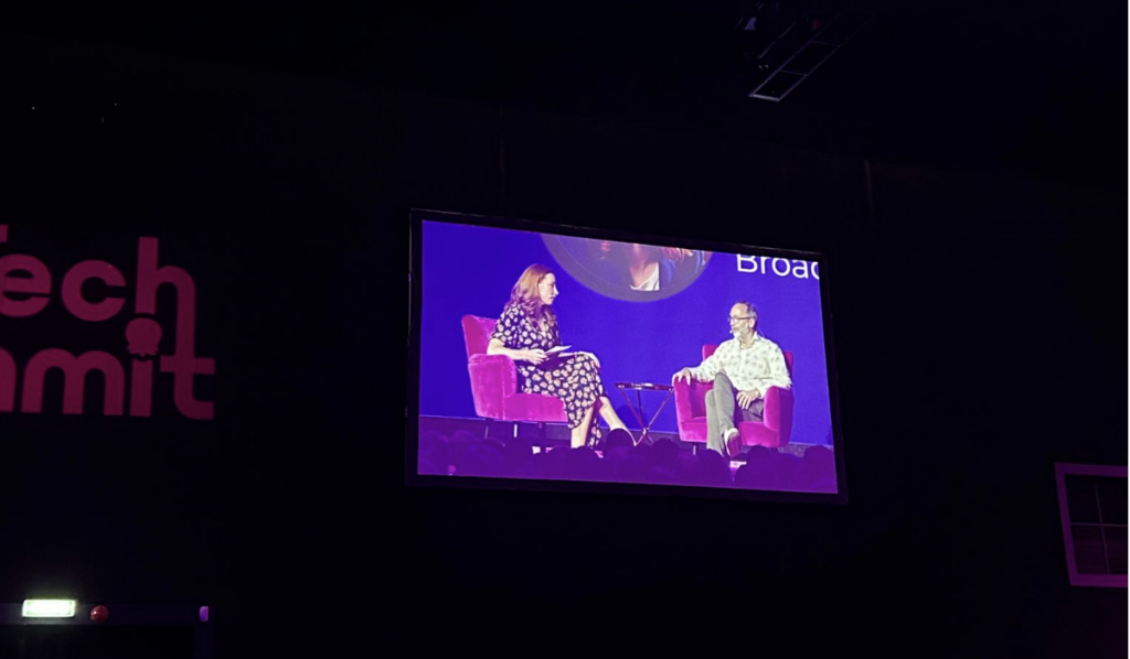 Side view of the audience at the Octopus Energy Tech Summit watching a large screen showing Professor Hannah Fry interviewing Wikipedia founder Jimmy Wales on stage. Purple lighting and a star-like ceiling create the conference atmosphere.