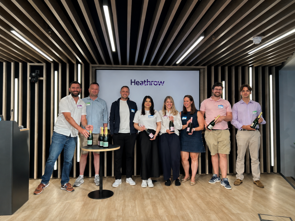 A group of eight smiling colleagues standing on a stage in front of a screen displaying the Heathrow logo, several holding bottles of champagne after winning a workshop challenge.